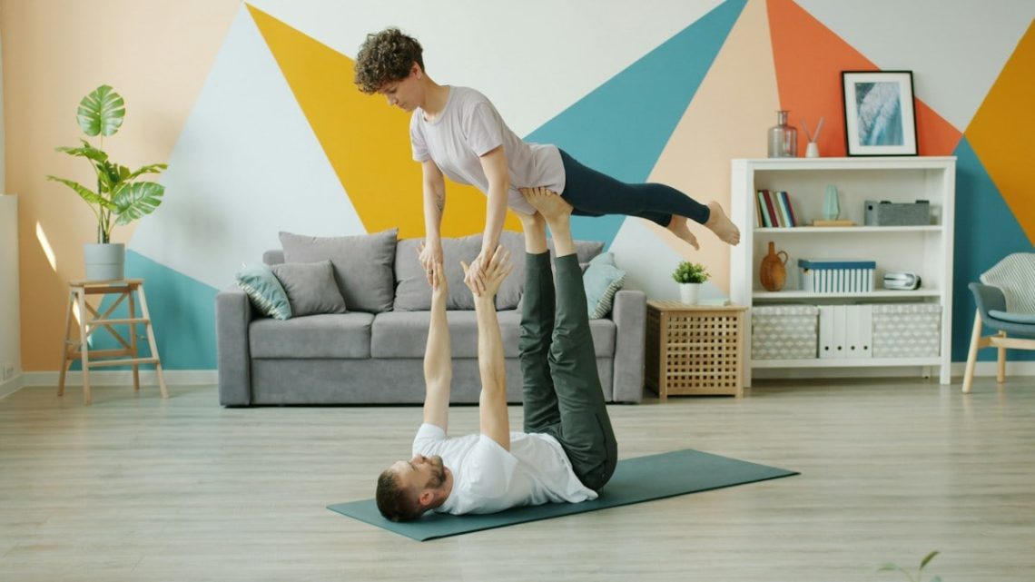 Happy couple is doing pair yoga at home, man is holding woman with legs on floor, girl is stretching arms. Relationship, active people and sports concept.