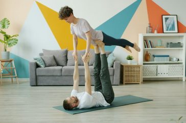 Happy couple is doing pair yoga at home, man is holding woman with legs on floor, girl is stretching arms. Relationship, active people and sports concept.