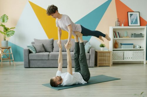 Happy couple is doing pair yoga at home, man is holding woman with legs on floor, girl is stretching arms. Relationship, active people and sports concept.